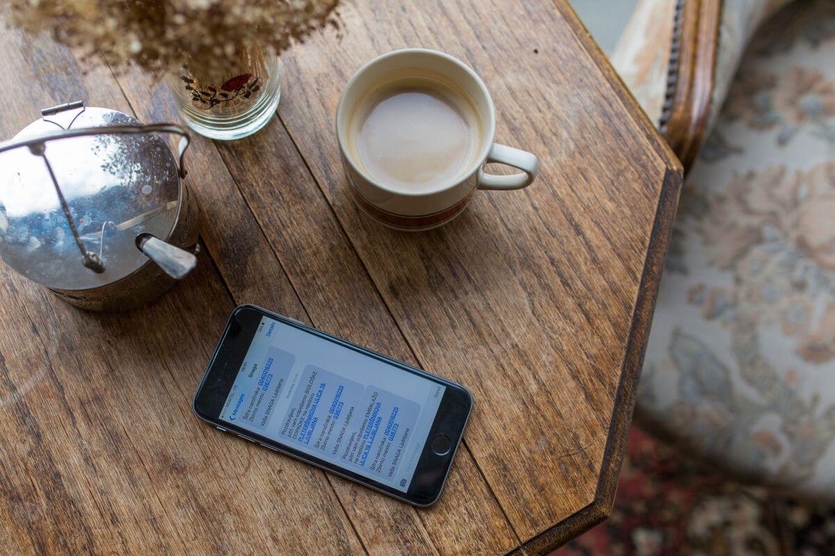 Miza, na kateri so mobilni telefon, na ekranu je prikazan SMS opomnik. Ob telefonu sladkornica, bela kava in vaza s suhim cvetjem.