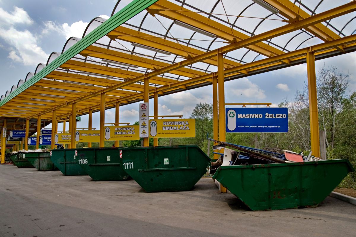 Večkubični zabojniki v Zbirnem centru Barje. Stojijo v vrsti, nad njimi so označevalne table in stojijo pod nadstrešnico.