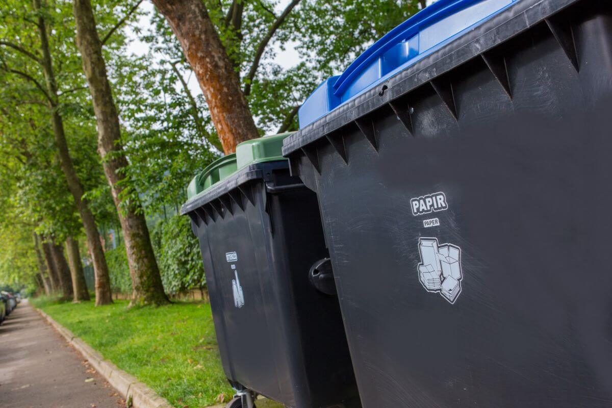 Štirikolesna zabojnika - v ospredju je zabojnik za papir, za njim zabojnik za steklo. Stojita na javni površini, ob cesti na eni strani in zelenico na drugi.