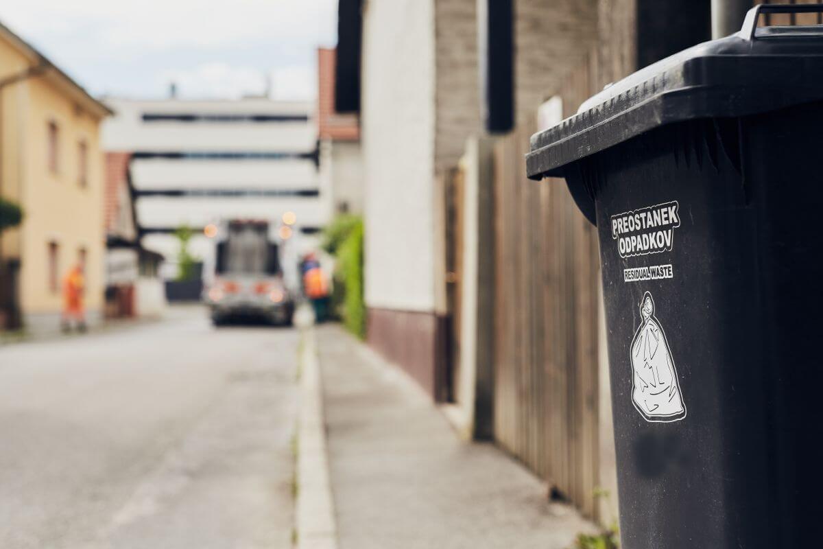 Črn zabojnik za mešane komunalne odpadke, v ozadju smetarsko tovorno vozilo in dva komunalna delavca.