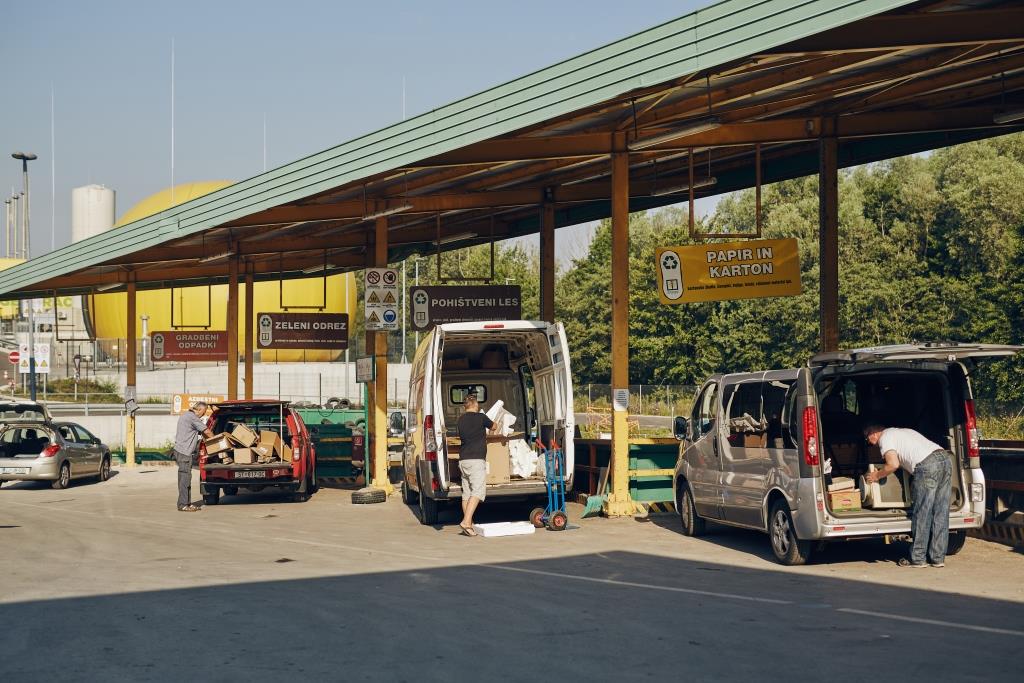 Trije občani iz odprtih prtljažnikov svojih vozil v zabojnike v Zbirnem centru barje odlagajo pripeljane odpadke. 