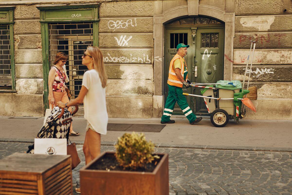 Ulični sprehajalci na ljubljanskih ulicah. Med njimi je tudi zaposleni Voke Snage z vozičkom za ročno čiščenje mestnih ulic.